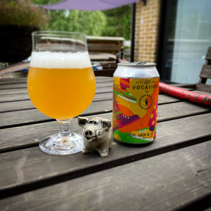 Ölglas fyllt med hazy ale, en ölburk och en liten grisskulptur på ett träbord utomhus.