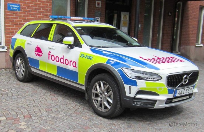 En bil med Foodoras färger och logotyp och blåljus parkerad framför en byggnad.