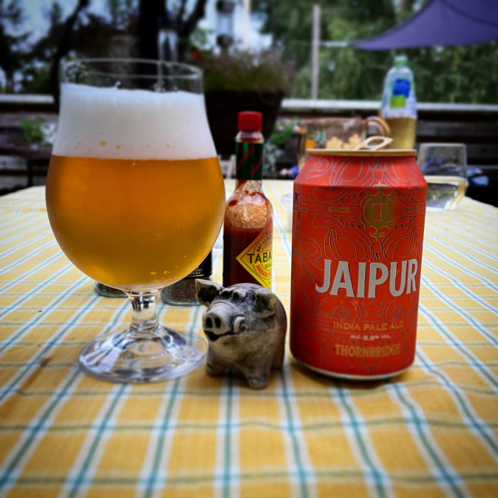 En ölglas fyllt med öl framför en "Jaipur India Pale Ale" burk med en Tabasco-flaska och en grisfigur på rutig duk.