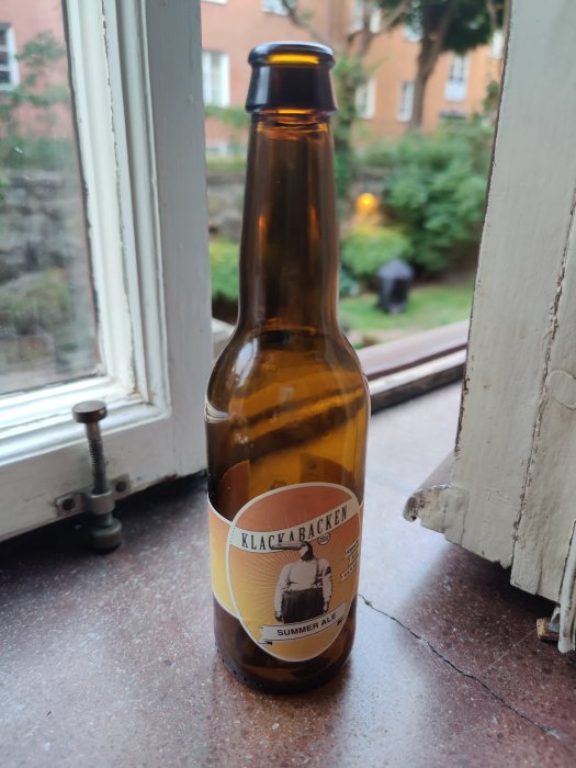Öppnad ölflaska av märket Klackabacken Summer Ale står på en fönsterbräda.