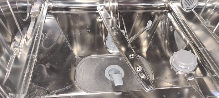 Insidan av en diskmaskin med metallkorgar, spolarmar och filter, klar för att laddas med disk.