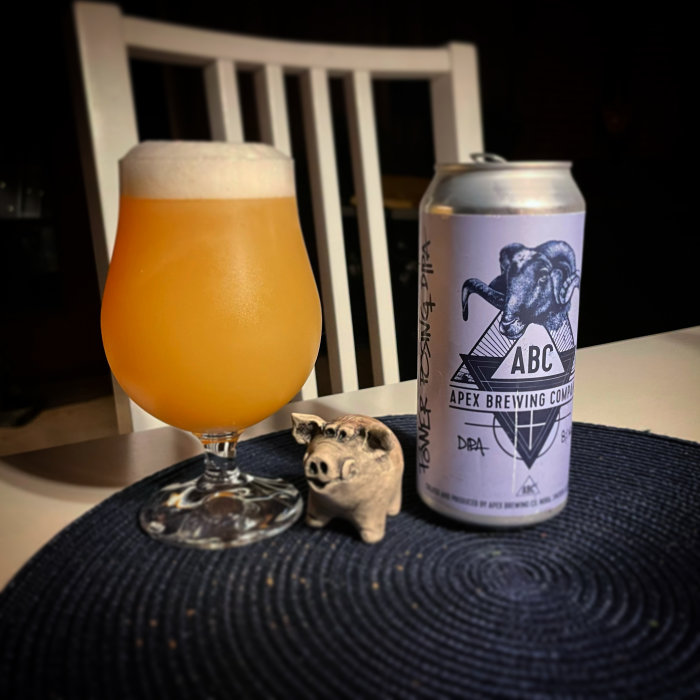 Ölglas fyllt med skummande öl bredvid en ölburk från Apex Brewing och en liten grisfigur på en mörk bordstablett.
