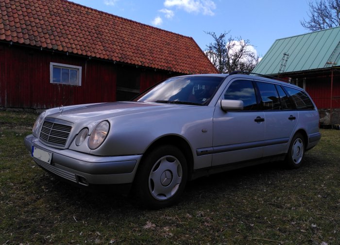 Silverfärgad Mercedes W210 kombi framför röda lador, i välbevarat skick utan synlig rost.