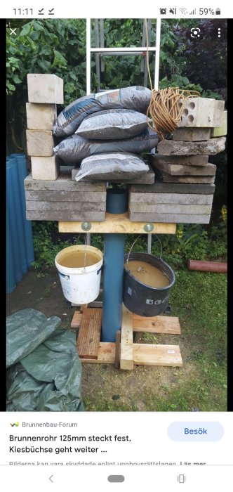 Improviserad stabilisering för ett brunnsrör med säckar, hinkar med vatten, plattor och träbitar på gräsmark.
