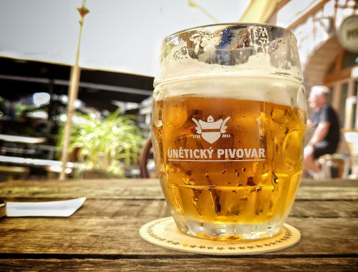 Halvfullt ölglas med logotyp på träbord, uteservering i bakgrunden.