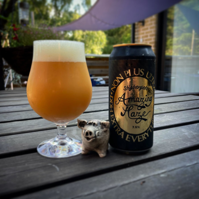 Ett ölglas fyllt med skummande öl bredvid en ölburk och en liten grisfigur på ett träbord.