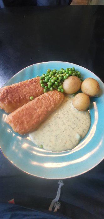Tallrik med två bitar stekt fisk, kokta potatisar, gröna ärtor och dillsås.