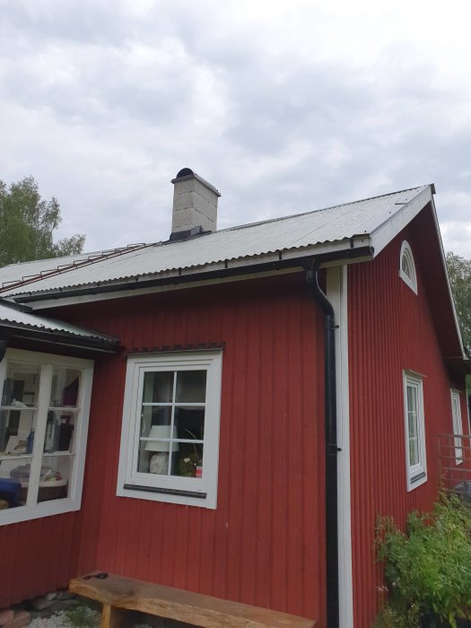 Rödmålat trähus med vita fönsterkarmar, plåttak och en murad skorsten syns under en molnig himmel.