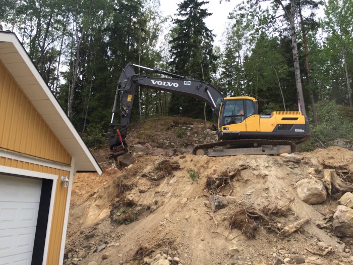 Volvo grävmaskin ränsar bergsida nära gul husfasad och vit garageport.