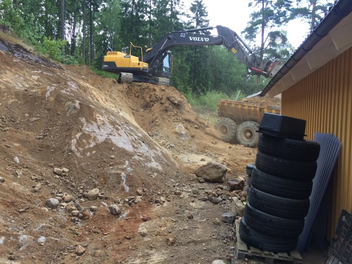 Grävmaskin på en byggarbetsplats flyttar jord medan en dumper väntar på last, arbete på en kuperad tomt.