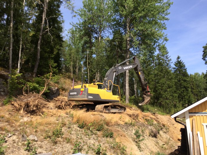 Grävmaskin vid arbete med att röja och jämna ut en bergssida, omgiven av träd och skog.