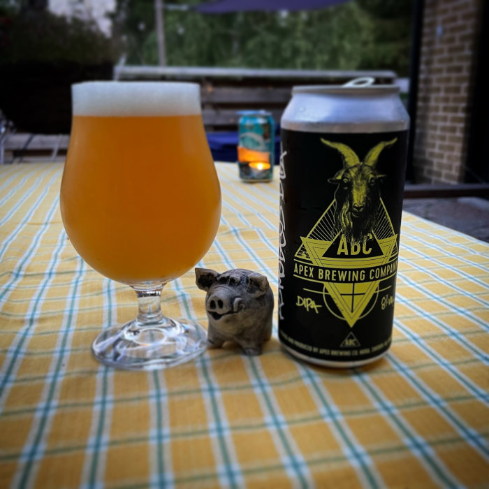 Ölglas fyllt med hazy IPA och en burk Apex Brewing DIPA på ett bord med små skulptur av en gris.