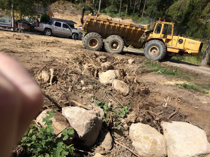 Gul 25-tons dumper som jämna ut tomten med utspridda stenar och rötter vid bergssida.