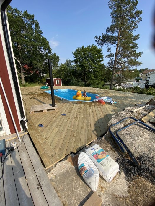 Nybyggd trädäck runt en ovanjordspool med uppblåsbara leksaker och byggmaterial i förgrunden.