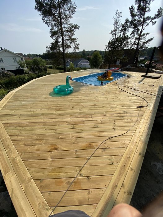 Nybyggd träterrass runt en utomhuspool med uppblåsbara leksaker, omgiven av natur och bostadshus.