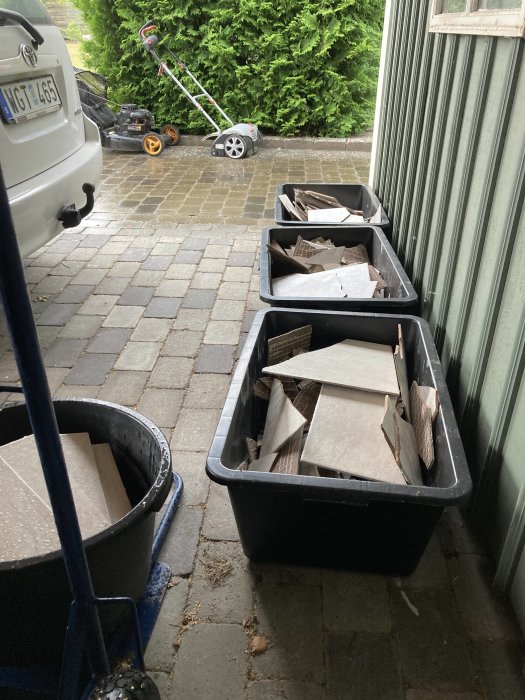 Klinkerplattor i plastlådor och en skottkärra i en carport, med en lövblås och en bil synlig utanför.