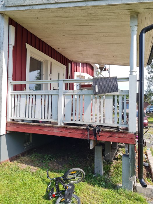 Altan fäst vid husets röda panel med vita staket, stöds av betongpelare, barnets cykel i förgrunden.