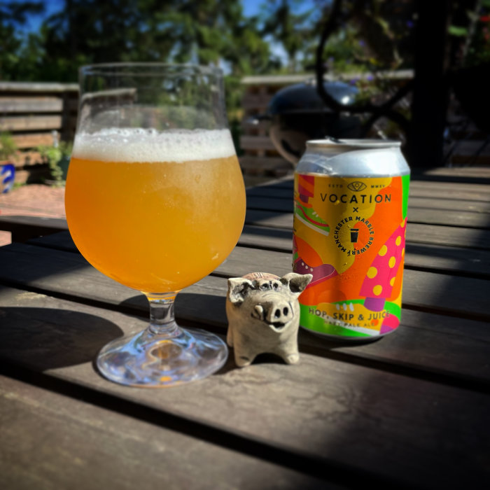 Ölglas med skum på en träbord bredvid en ölburk och en liten prydnadsgris, utomhus i soligt väder.