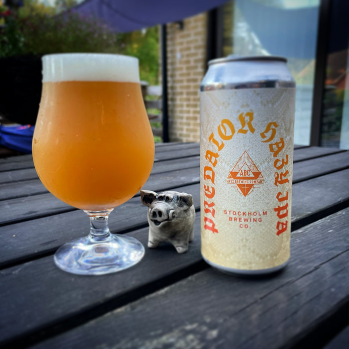 Ölglas fyllt med grumlig dryck bredvid en burk från Stockholm Brewing Co. på ett utomhusbord, liten grisfigur i förgrunden.