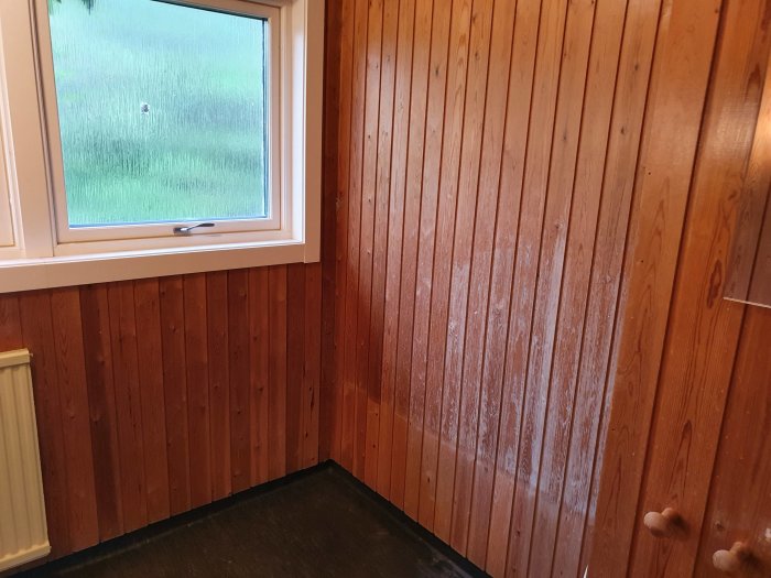 Ett badrums hörn med brunt träpanel och ett fönster, planerat för badkar nära fönstret enligt GVK-krav.