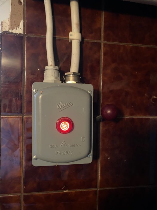 Elektrisk säkerhetsbrytare med lysande röd indikatorlampa, omgiven av kakelvägg.