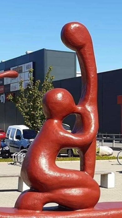 Abstrakt röd skulptur utanför shoppingcenter under klarblå himmel.