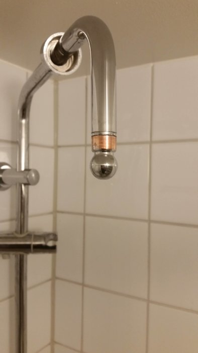 Duscharm i krom fast i väggen ovanför vita kakelplattor, med kalkavlagringar vid anslutningen.