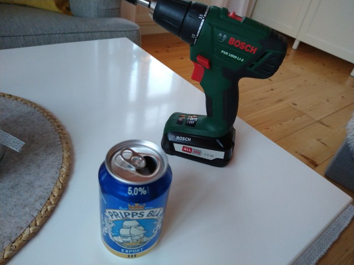 En öppnad burk Pripps Blå öl bredvid en grön Bosch skruvdragare på ett vitt bord.