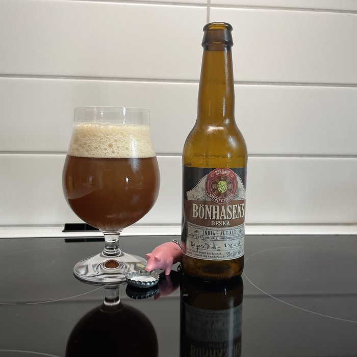 Öppnad ölflaska och ett glas med skummande öl på glänsande bord, med en kapsyl och en liten rosa figur.