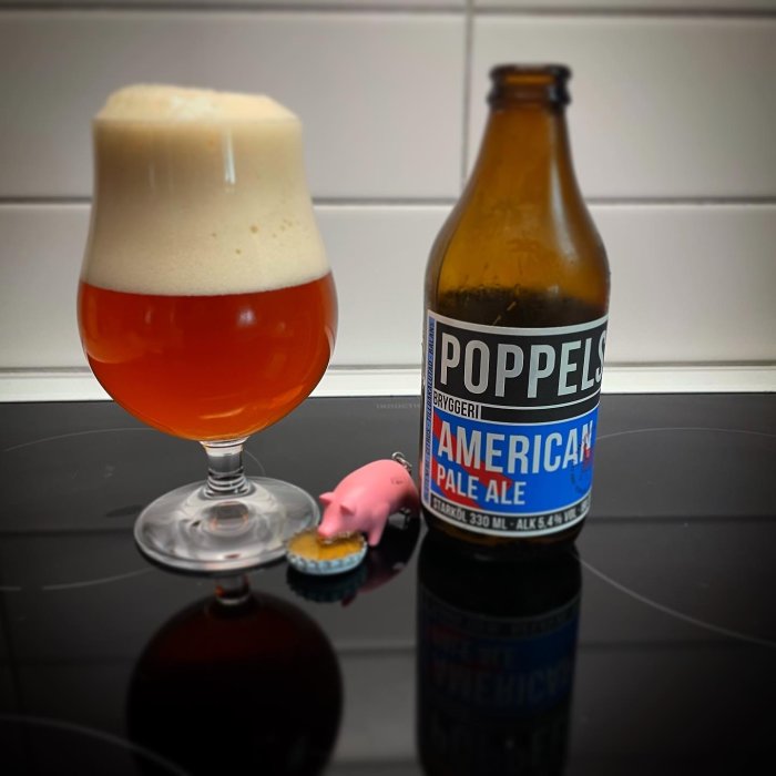 Öppnad Poppels American Pale Ale-flaska bredvid ett glas öl med tjockt skum, och en liten rosa plastgris på en kapsyl.