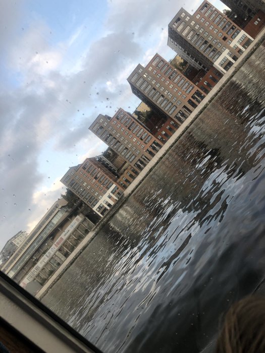 Vy genom fönster med vattendroppar på glaset med utsikt över en flod och moderna byggnader i skymningen.