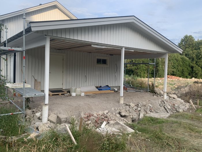 Carport under konstruktion vid ett hus med grovplanerad mark och byggmaterial synliga.