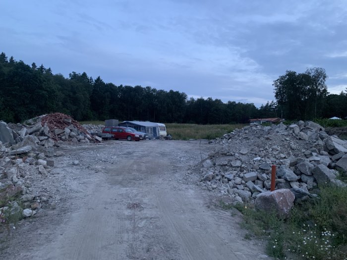 Grovplanering av en tomt med hjullastare, stora grushögar, röd bil i carport och skog i bakgrunden.