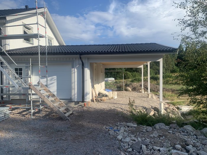 Nybyggd carport med pågående markplanering och grusfyllning vid vit husfasad.