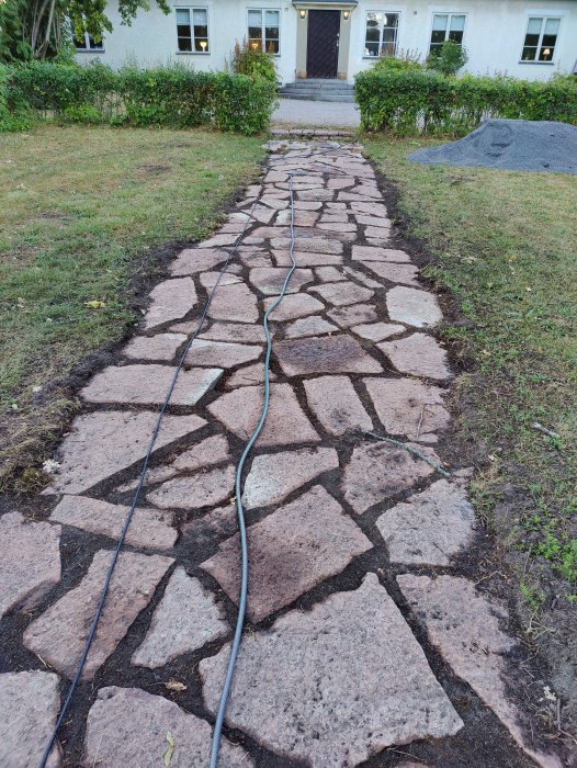 Stenbelagd gångväg med ojämna stenar och jord mellan stenarna som leder till ett hus.