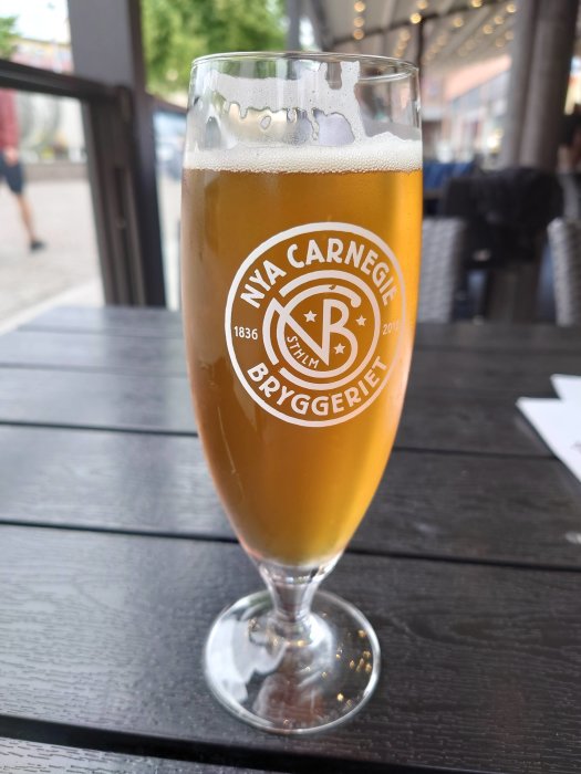 Ett glas ljust öl med Nya Carnegiebryggeriet-logga på ett uteserveringsbord.