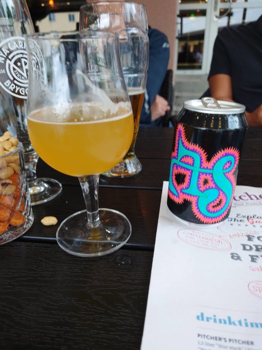 Ölglas halvfyllt med grumlig öl och en färgrik ölburk på ett bord med meny, indikerar en provsmakning av en lokal NEIPA och en ny Omnipollo öl.
