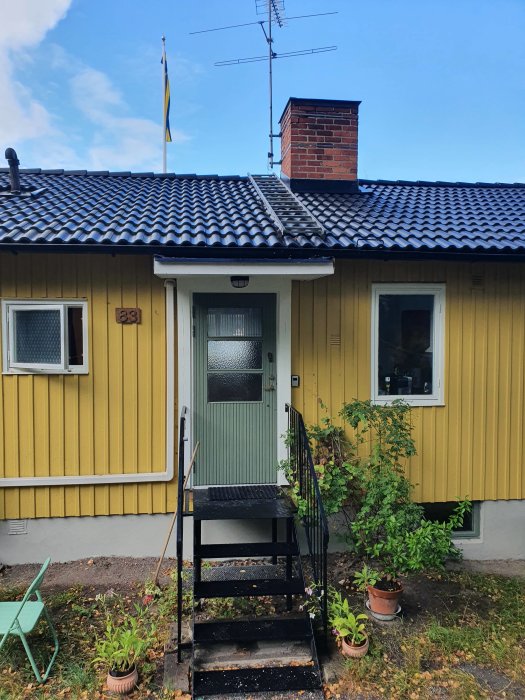 Entré till ett gulmålat hus med svarttak och vindskiveplåt, svensk flagga i bakgrunden, trappa och gröna växter framför.