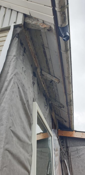 Slitet takfot med flagande färg och synliga träbrädor vid ett fasadbytprojekt, täckt med byggfolie och ställningskomponenter.