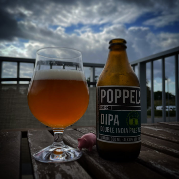 Ölglas och Poppels bryggeri DIPA-flaska på ett träbord med molnig himmel i bakgrunden.