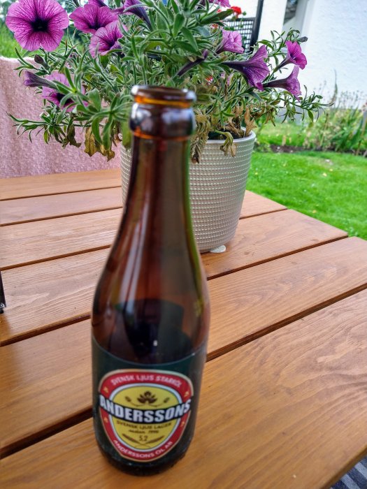 Öppnad grön ölflaska "Anderssons Lager" på träbord framför kruka med blommor.
