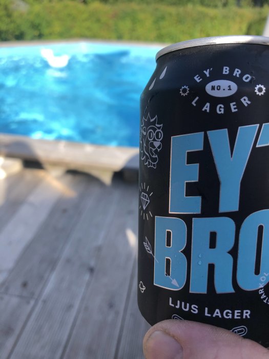 En burk lageröl hålls i förgrunden med en otydlig bakgrund av en pool och ett trädäck.