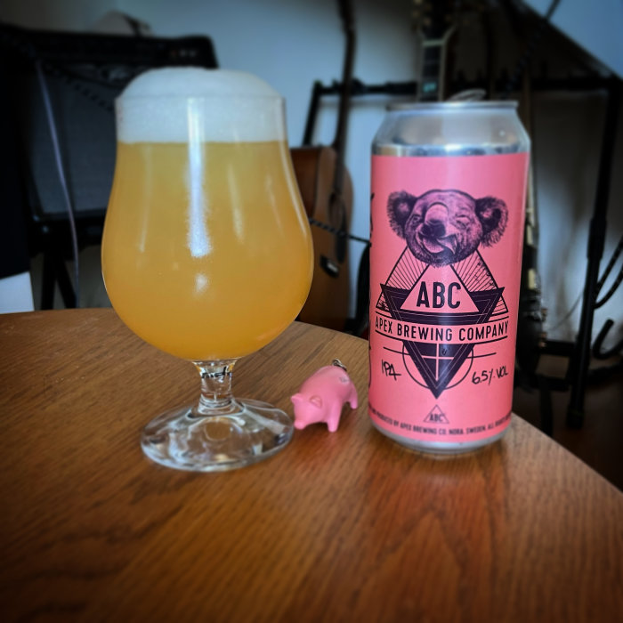 Ölglas fyllt med öl bredvid en ölburk och en liten plastgris på ett träbord.