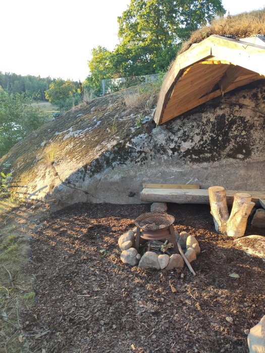 En nybyggd grillplats med stenar runt, grillgaller och bänk, under visande berg och träd.