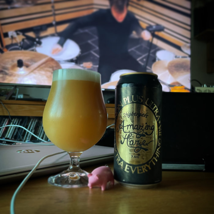 Ölglas med skum på ett skrivbord, burk med etikett, liten plastgris, datorskärm med trumslagare i bakgrunden.