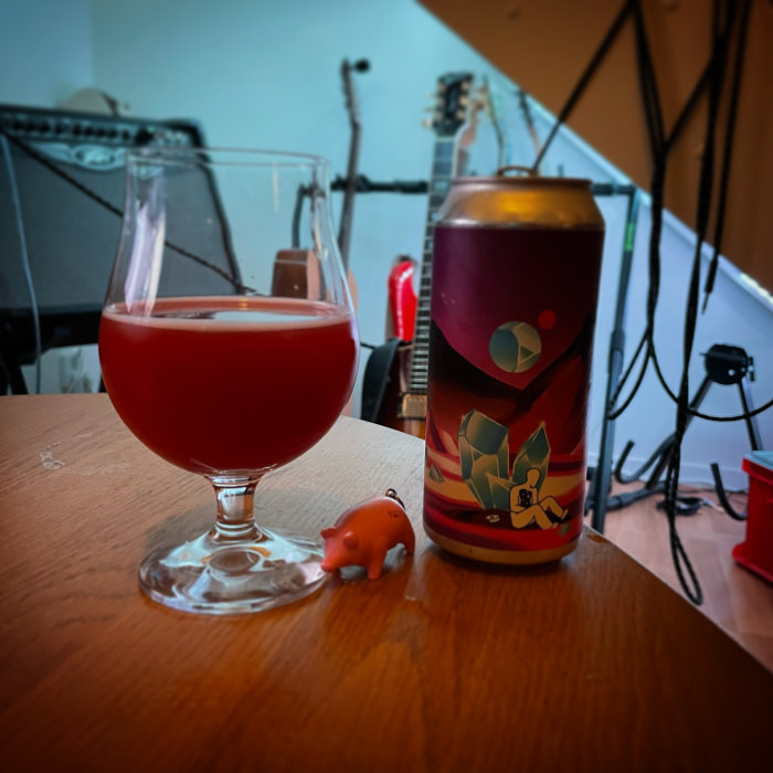 Ett glas med röd dryck bredvid en färgglad ölburk och en liten grisknorrfigur på ett bord framför gitarrer.