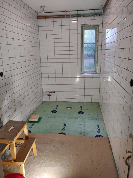 Nyligen kaklat badrum med vita plattor, ett fönster med kakellist, och oulagt golv.