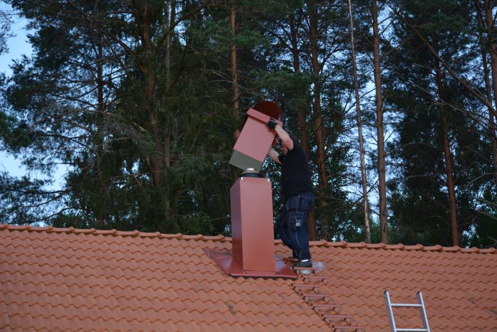 Person installerar skorsten på taket av ett hus omgivet av träd i skymningen.