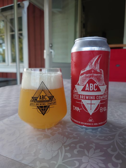 Ölglas fyllt med öl bredvid en burk från Apex Brewing Company på ett bord.