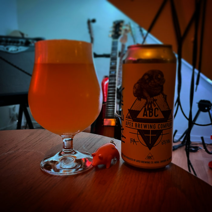 Ett glas med orange dryck och en ölburk från Apex Brewing bredvid en liten gummigris på ett bord.